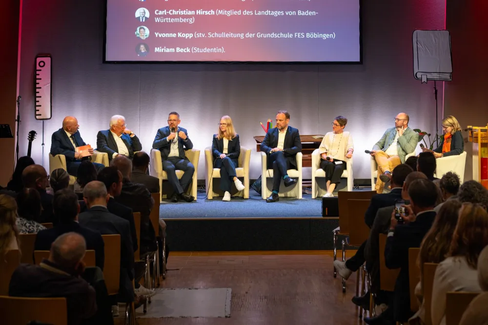 Podiumsdiskussion auf der Bühne: acht Personen in hellen Sesseln, über ihnen eine Leinwand mit den Teilnehmerinnen und Teilnehmern