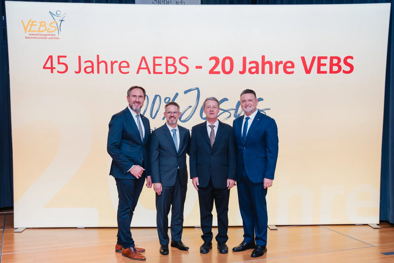 Vier Männer in dunklen Anzügen vor dem Banner 45 Jahre AEBS – 20 Jahre VEBS