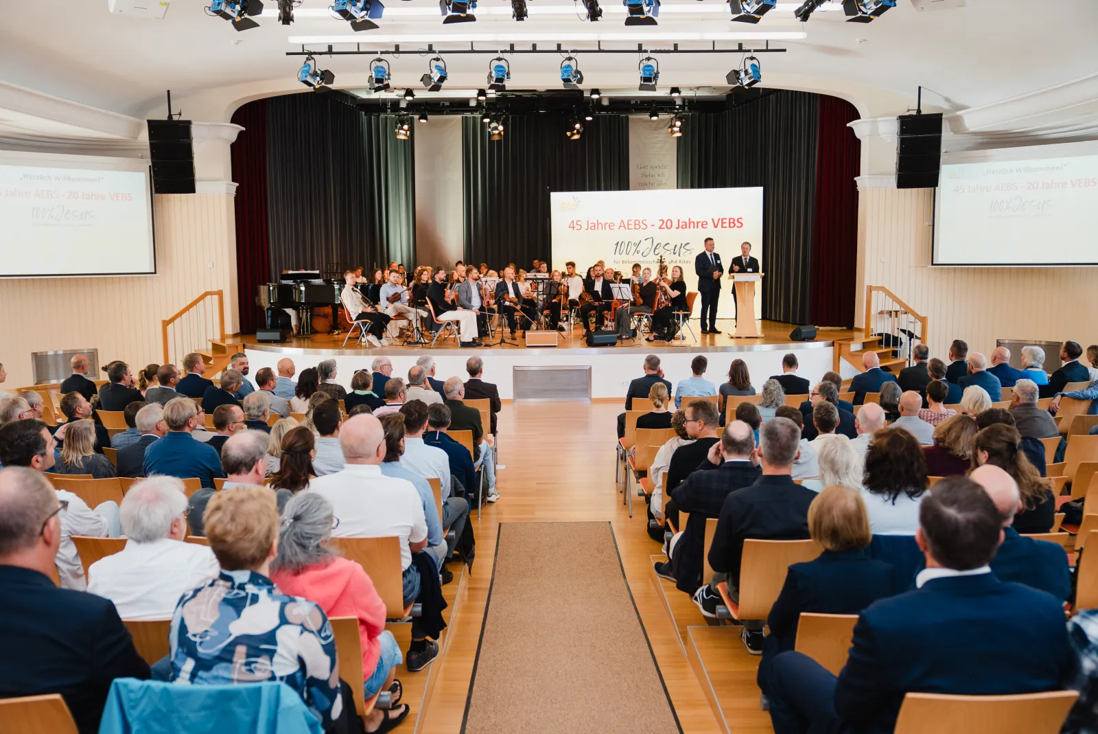 Blick in den Festsaal in Detmold: Gäste im Vordergrund, auf der Bühne Orchester und Rednerpult vor dem Banner 45 Jahre AEBS – 20 Jahre VEBS