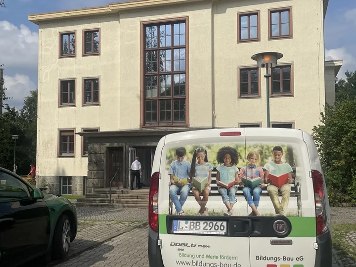 Mehrstöckiges helles Schulgebäude, davor ein Kleinbus mit einem Foto lesender Kinder